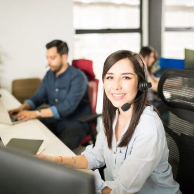 Portrait of pretty young latin woman wearing a headset sitting at her office desk with coworkers in background